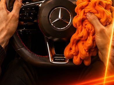 Hands stretching orange kinetic dough off a Mercedes-AMG steering wheel